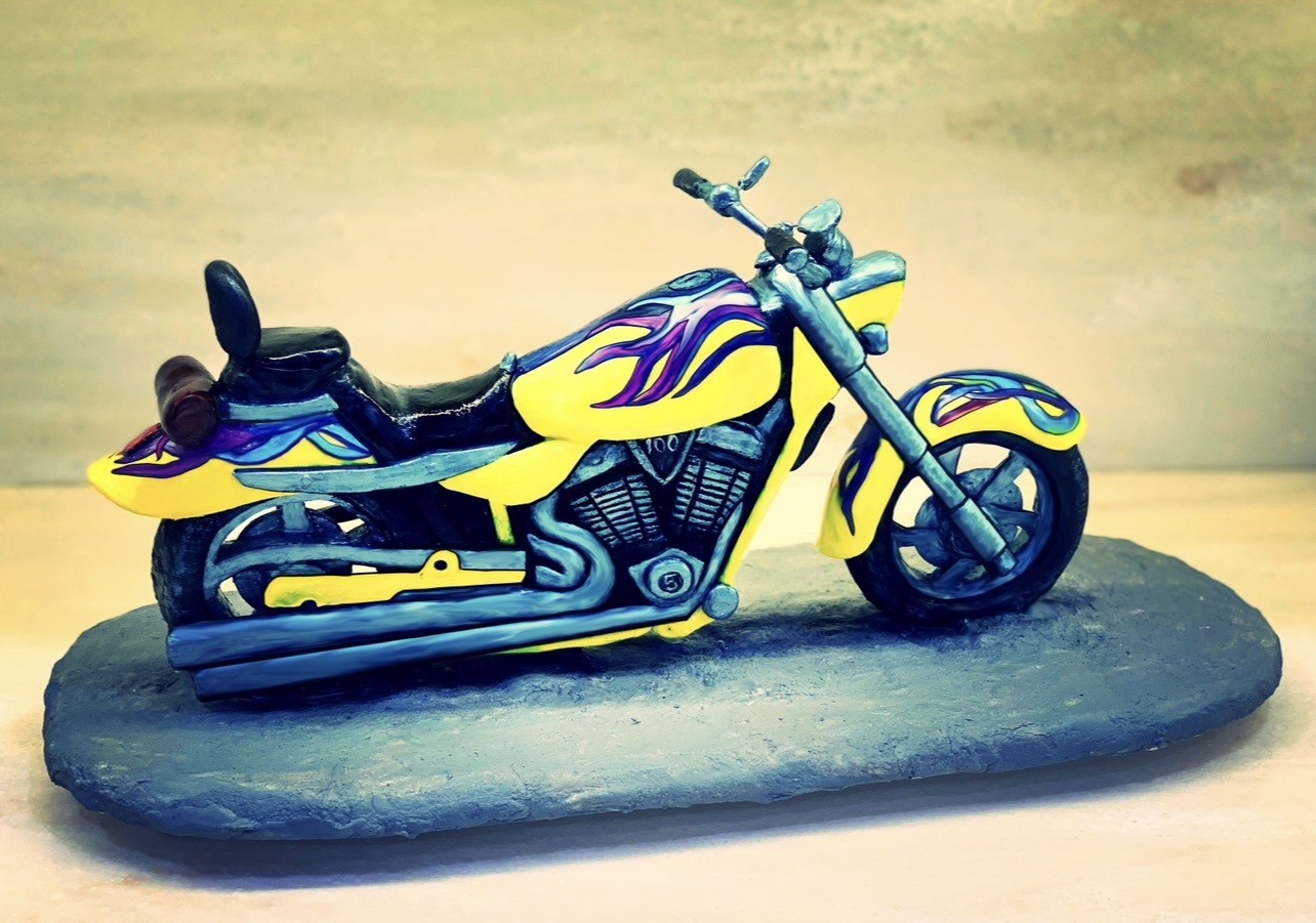 A sculpted cake of a custom chopper-style motorcycle mounted on textured slate-gray base resembling rough stone, electric yellow body panels with purple and blue flame decals, metallic gray engine and exhaust pipes, black seat and rear fender, "100" emblem on tank.