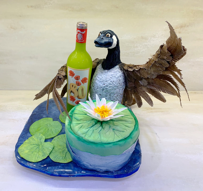 An elaborate cake decorated to resemble a pond scene features a sculpted goose with spread wings standing behind a round cake topped with a water lily, accompanied by a green fondant bottle marked with the number 80.