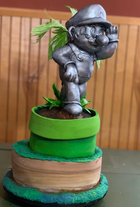 Super Mario cake with a Mario figurine emerging from a green pipe on a tiered dirt-mound base.