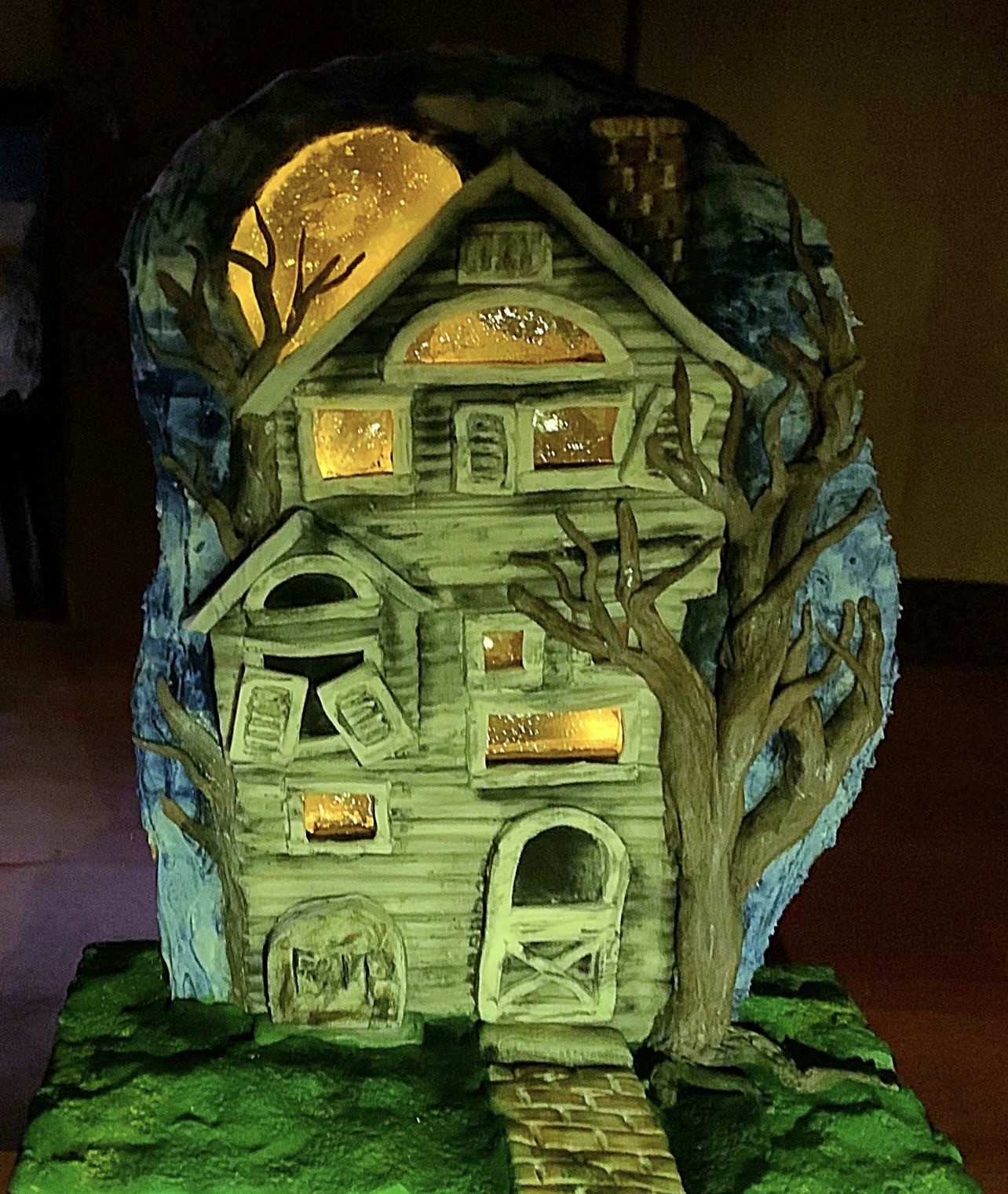 A haunting sculpted cake diorama of a dilapidated Victorian-style house with asymmetrical windows aglow with amber light, an arched "X"-braced doorway, weathered stone marker, mossy green terrain with cobblestone path, surrounded by gnarled leafless trees under a luminous full moon in warm golds and yellows.