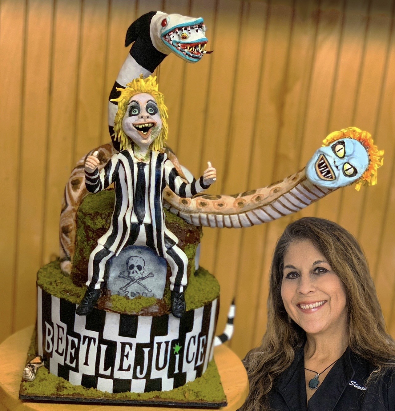 A sculpted cake tribute to Beetlejuice featuring the character in his iconic striped suit with spiky yellow hair and mischievous grin, perched on a moss-covered tombstone with skull and crossbones, two serpentine creatures emerging from behind — one menacing, one comical — on a black-and-white checkered base with bold "BEETLEJUICE" lettering.