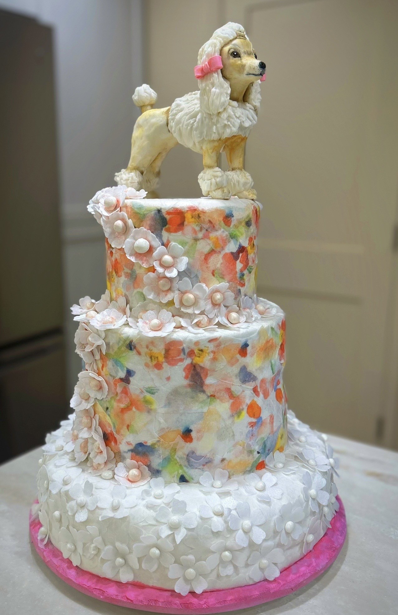A three-tiered custom cake with a sculpted poodle figurine topper featuring textured white fur, golden-tan accents on legs and face, and a pink bow on its ear, the top two tiers wrapped in vibrant watercolor-style fondant with soft washes of coral, peach, lavender, mint, and sky blue, adorned with cascading sugar flowers.