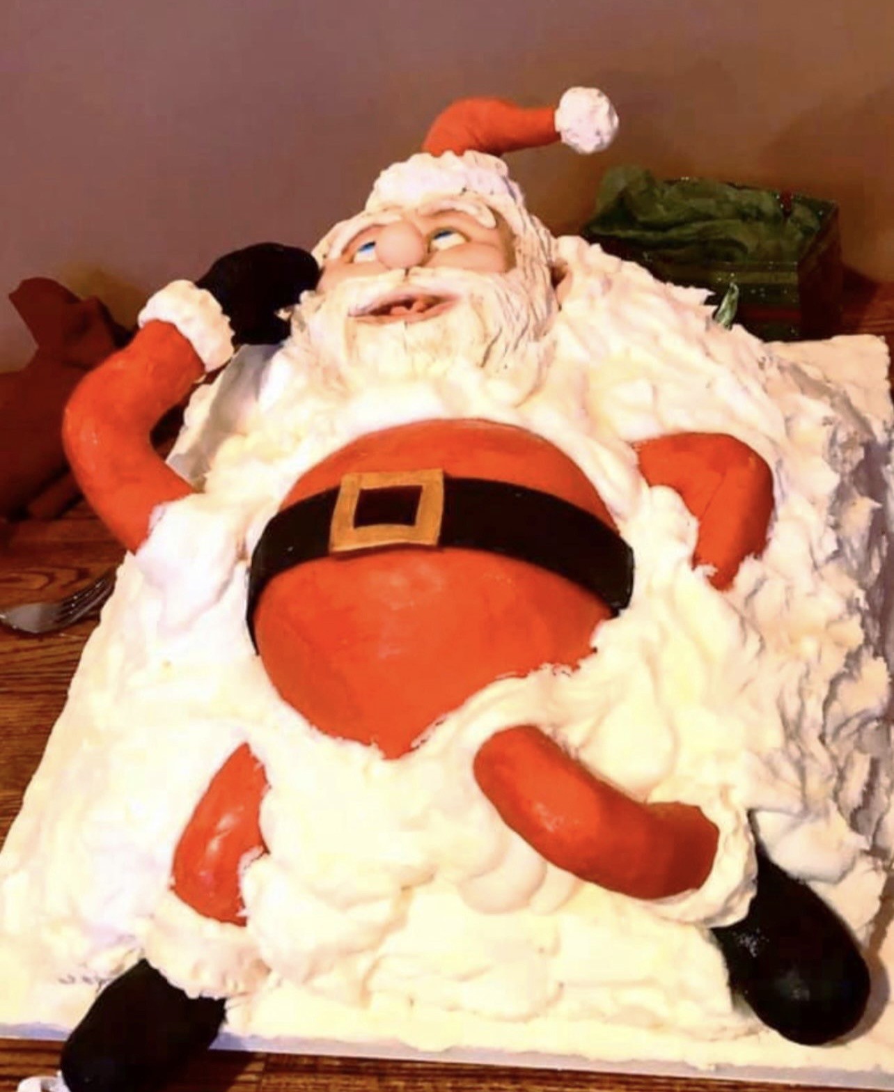 A whimsical sculpted cake of Santa Claus reclining lazily on his back atop fluffy white frosting mimicking snow, his body exaggeratedly round in bright red fondant suit with black belt and boots and gold buckle, expressive blue eyes, rosy cheeks, wide goofy grin, one hand behind his head.
