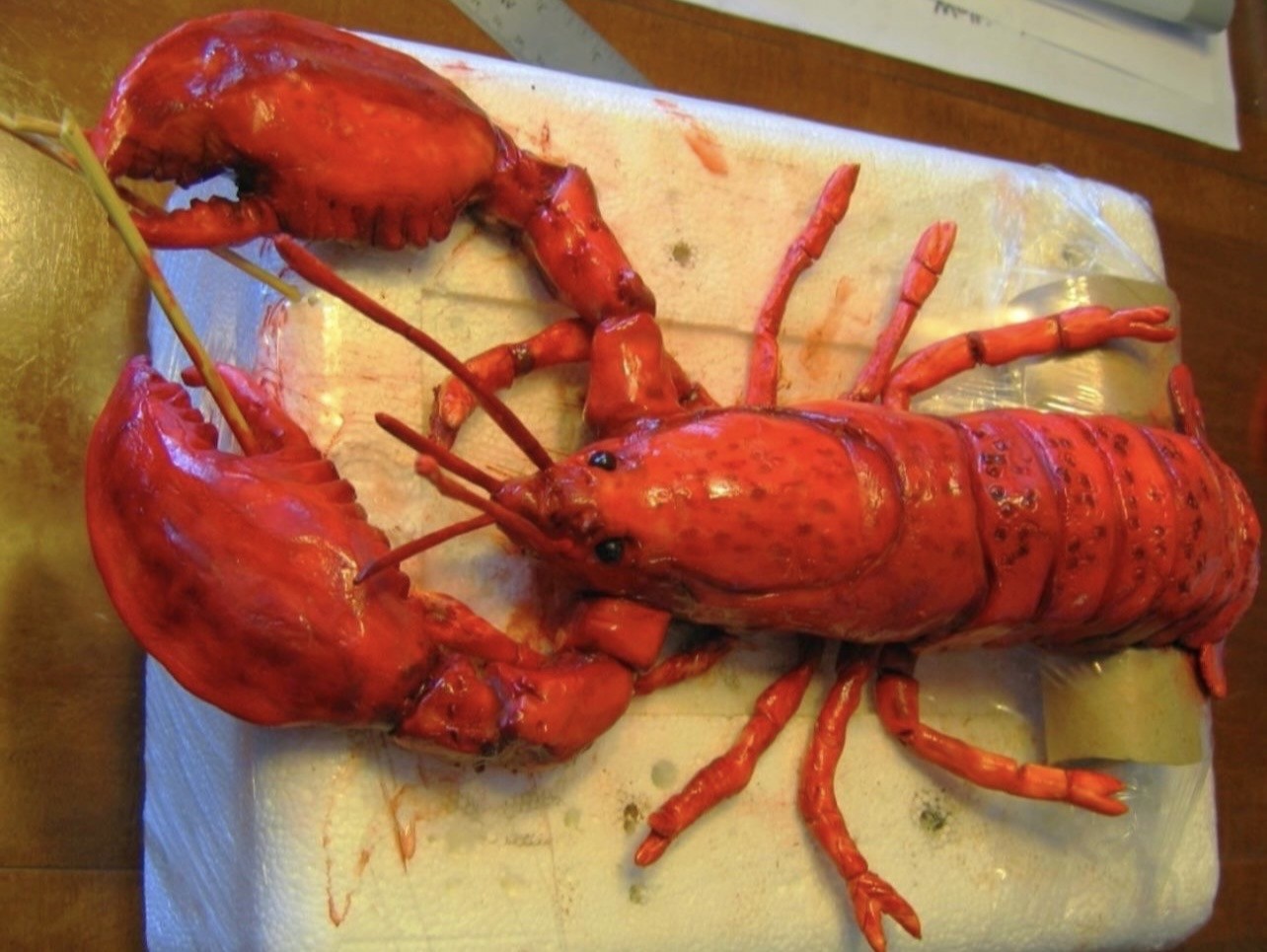 A hyper-realistic sculpted cake of a cooked lobster in vivid glossy red tones with segmented tail, articulated legs, textured carapace, and tiny black eyes, positioned on a white block with wooden skewers piercing through the claws.