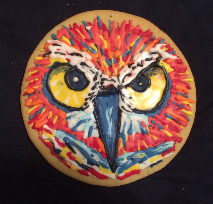 A round cookie decorated with colorful icing to resemble an owl's face, featuring large yellow eyes, a blue beak, and vibrant red, orange, and blue feather patterns.