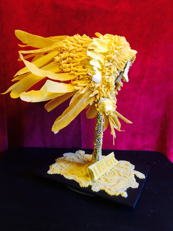 A sculpted sunflower resembling flower with a stem made of small pasta beads and a base of lace-like pasta material, featuring the word "LIBERTY" on a small sign at its base, set against a vibrant red background.