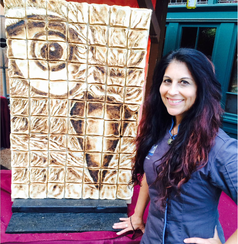 A smiling woman with long dark hair stands next to a large portrait of Albert Einstein created from a grid of toasted marshmallows arranged in pixelated form.