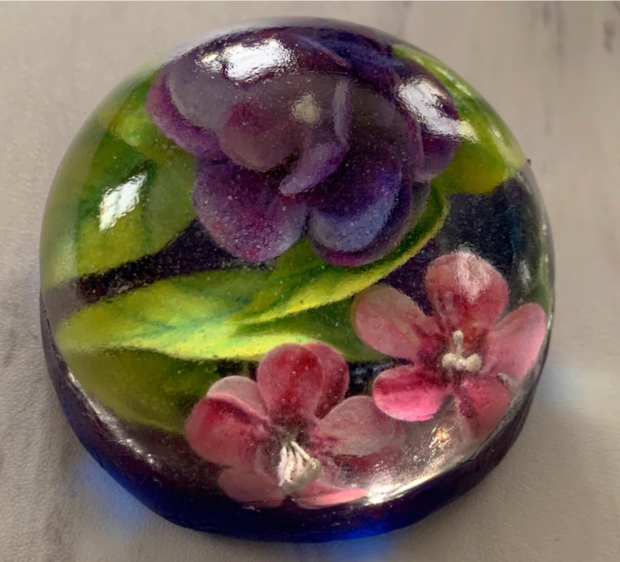A sugar art glass-like paperweight features a floral design with purple and pink sugar blossoms and green leaves set against a dark background.