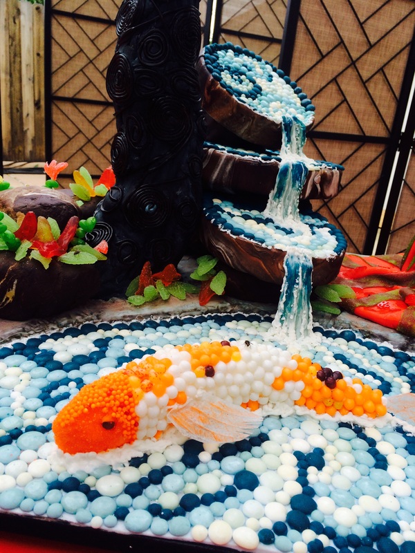 An elaborate sugar art piece designed as a koi pond features a waterfall cascading down chocolate rocks into a pool of blue and white candies, where a large koi fish made of orange and white sprinkles swims.
