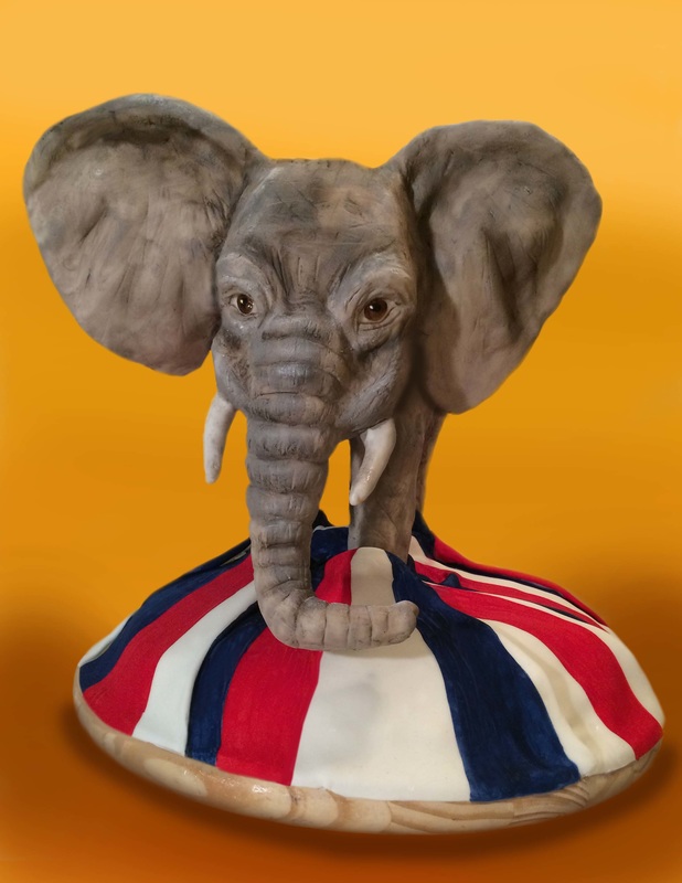 An elephant-shaped cake with large ears and white tusks emerges from a round base decorated with red, white, and blue stripes, set against a solid orange background.