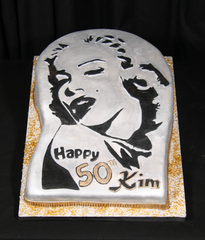 A silver-frosted cake features a black-and-white stencil of Marilyn Monroe with "Happy 50th Kim" written in black and gold icing, set against a black background with gold sprinkles on the white cake board.