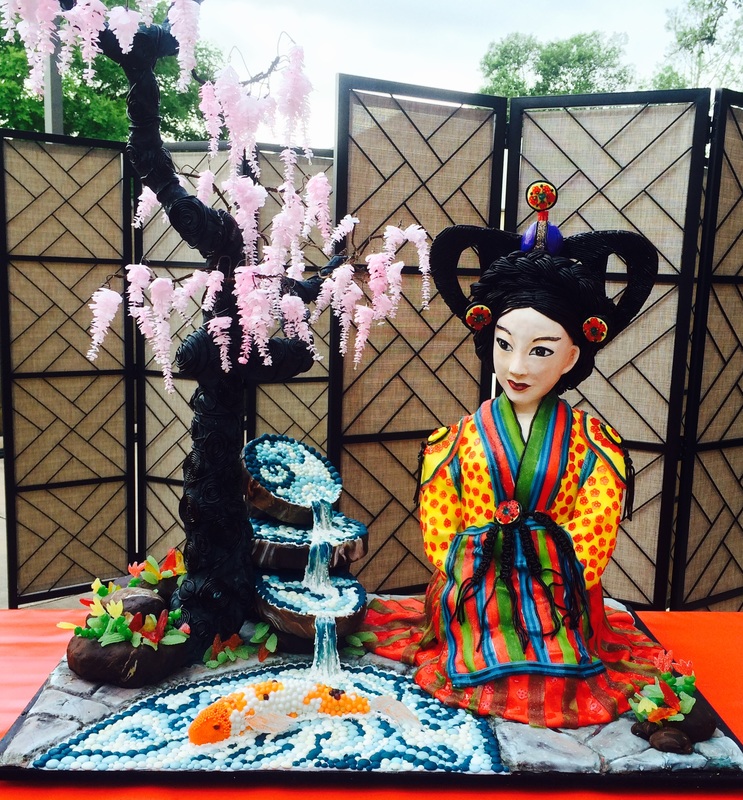 An elaborate sugar art sculpture features a geisha figure in a colorful kimono seated beside a black wisteria tree and a cascading waterfall with koi fish, all set against a traditional Japanese folding screen.