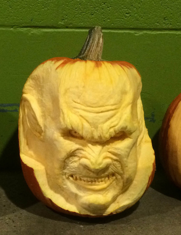 A carved vegetable features a detailed, menacing face with a furrowed brow, sharp teeth, and pointed ears, set against a green wall.