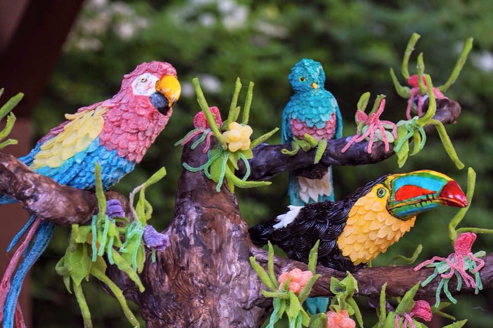 A colorful sugar sculpture of three tropical birds, including a macaw, a toucan, and a smaller parrot, perched on a branch adorned with green leaves and pink flowers.