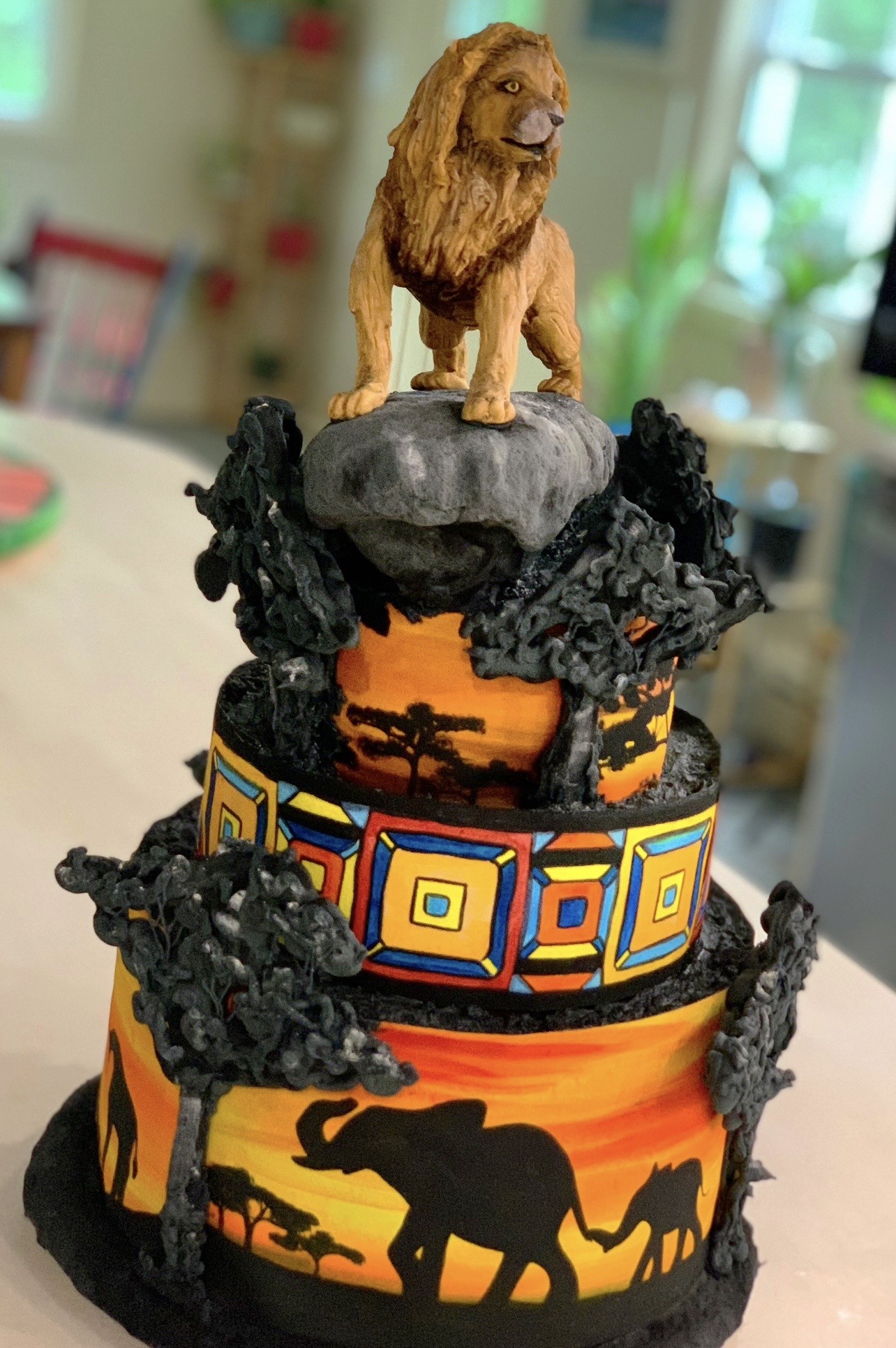 A two-tiered African savanna-themed cake with a sculpted lion figurine atop a gray fondant rock, vibrant sunset backdrops in orange and yellow with acacia tree silhouettes, Kente-inspired geometric patterns in red, blue, yellow, and black, black fondant elephant and giraffe silhouettes against the glowing sky, and dark fondant bushes flanking both tiers.