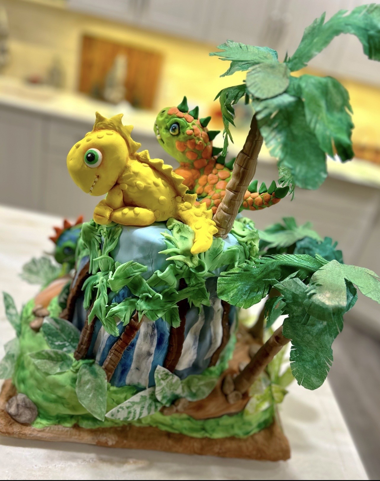 A dinosaur-themed cake featuring a multi-tiered prehistoric island with vibrant green foliage, palm trees, and rocky terrain, topped by two hand-sculpted fondant dinosaurs — a bright yellow one in the foreground and a green-and-orange one behind — with expressive eyes and playful expressions.