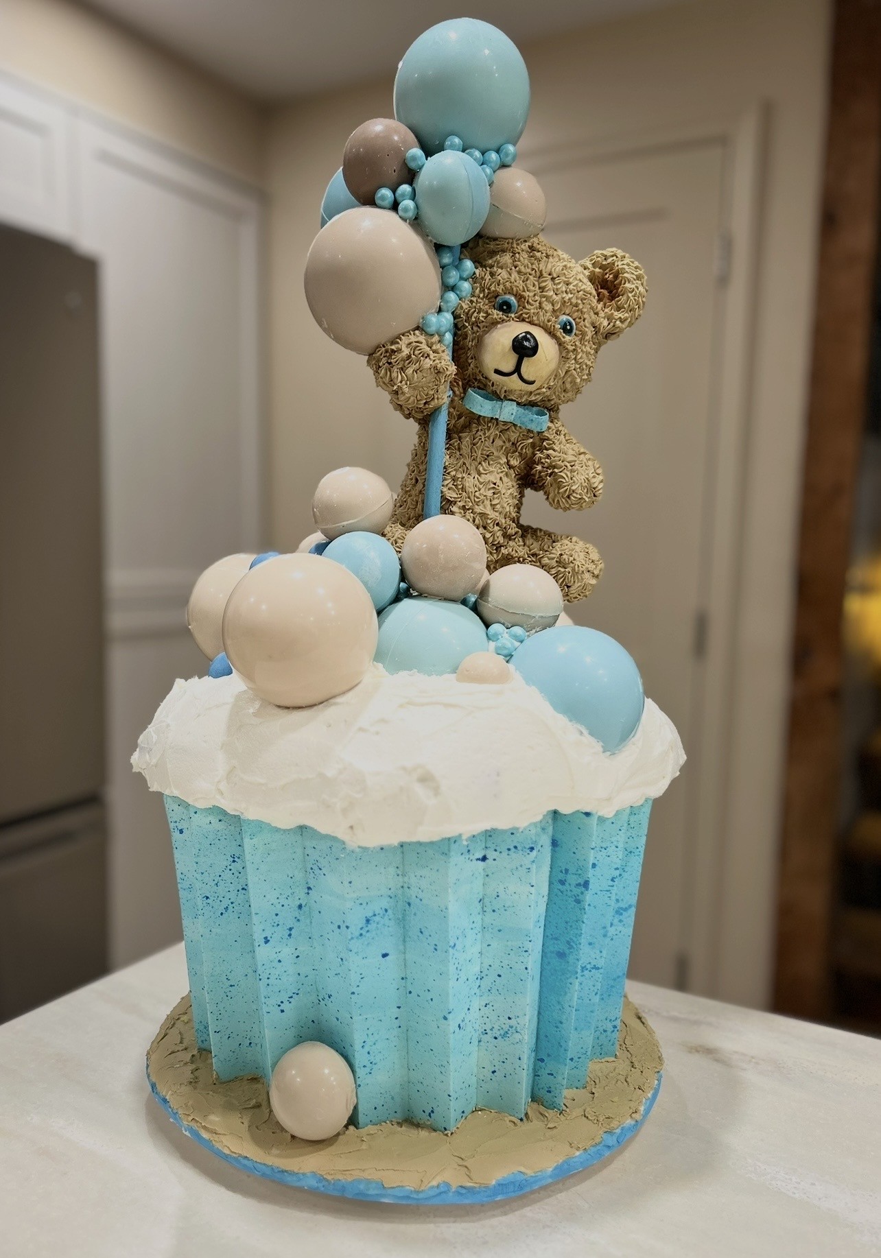 A cupcake cake designed as an oversized blue cupcake liner with white frosting, decorated with pastel-colored spherical candies, topped by a meticulously sculpted teddy bear in tan fondant with a light-blue bow tie holding a cluster of glossy balloon-like spheres.