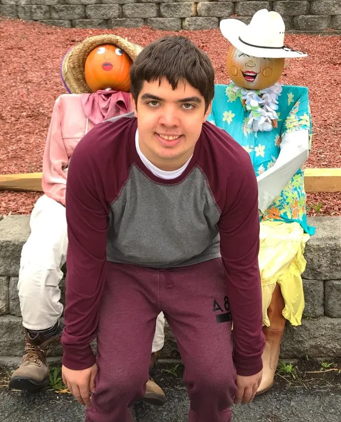 Devin Michael Housman smiling in a maroon and gray sweatshirt kneeling by scarecrows.