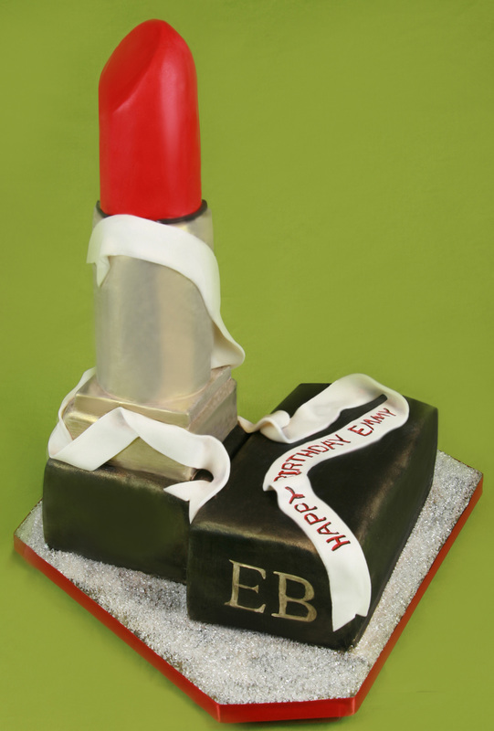 A lipstick-shaped cake with a red top and silver casing sits upright next to its black case, which is decorated with a white ribbon reading "Happy Birthday Emmy" and the initials "EB."