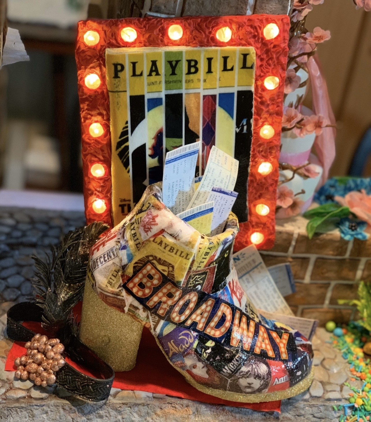 A Broadway-themed sculpted cake featuring a high-heeled platform pump covered in decoupage of Playbills, ticket stubs, and bold "BROADWAY" marquee lettering, heel and toe dipped in shimmering gold glitter, accompanied by a rectangular frame wrapped in crinkled red tissue paper illuminated by warm LED lights with a stylized Playbill cover.