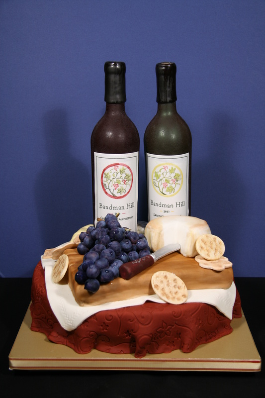 A sculpted cake designed to look like a wine and cheese platter, featuring two fondant wine bottles labeled "Bandman Hill," a cluster of purple grapes, a wedge of cheese, crackers, and a knife, all arranged on a wooden board atop a red tablecloth.