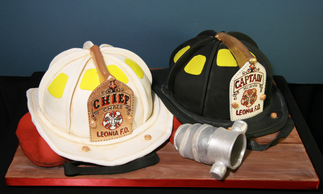 Two elaborate fondant cakes shaped like firefighter helmets, one white labeled "Chief" and one black labeled "Captain," sit on a wooden board alongside a silver cake replica of a fire hydrant.