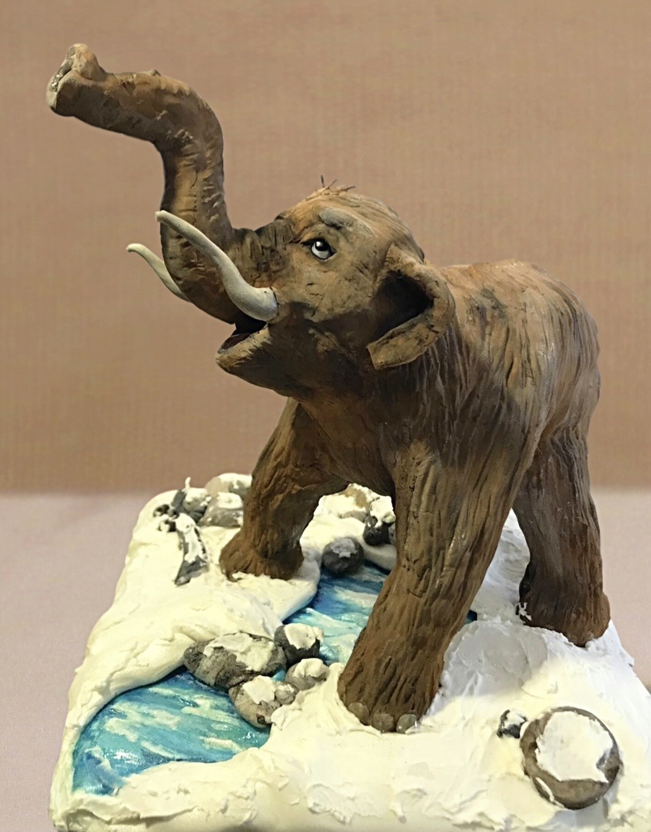 A sculpted cake of a woolly mammoth captured mid-stride with trunk raised and curled, curved ivory tusks contrasting with rich textured brown fur showing fine striations mimicking real hair, glossy black eye with white highlight on a diorama base designed as snowy icy landscape with white snow drifts, gray rocks dusted with faux snow, and winding blue frozen river.