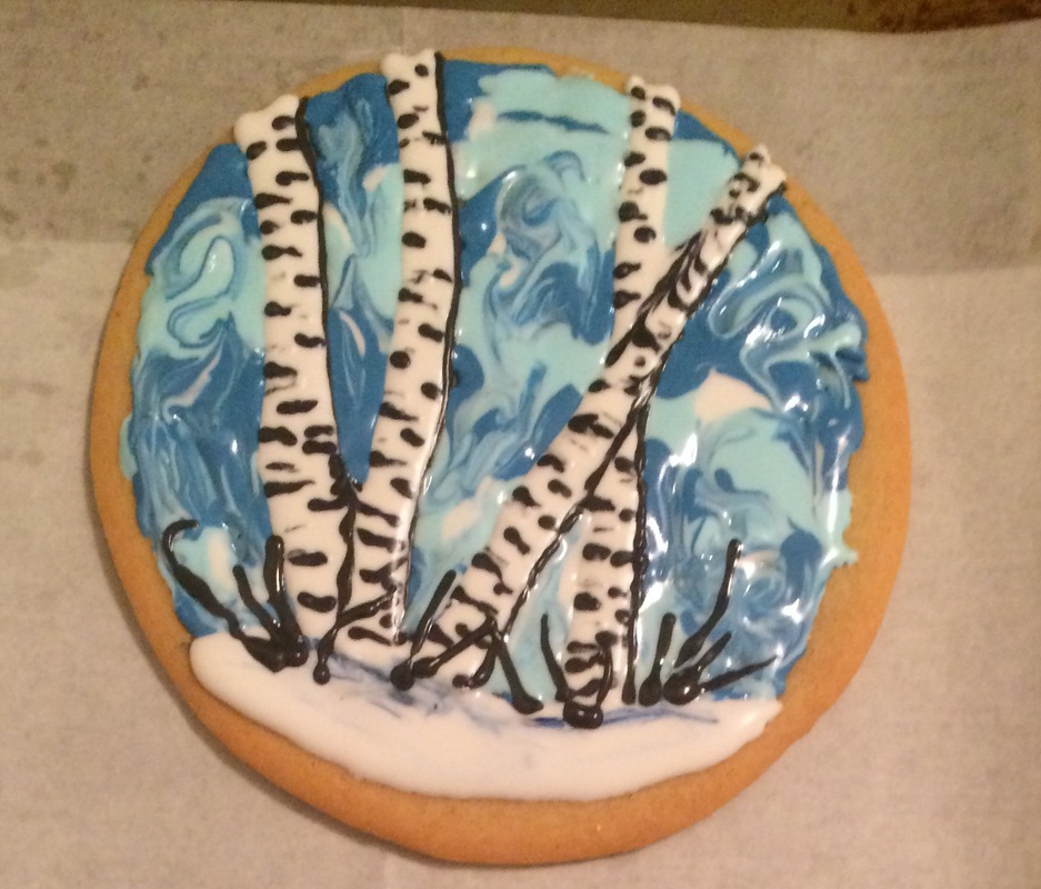 A round sugar cookie decorated with royal icing to depict a winter scene featuring white birch trees with black markings against a swirled blue background and a white snowy base.