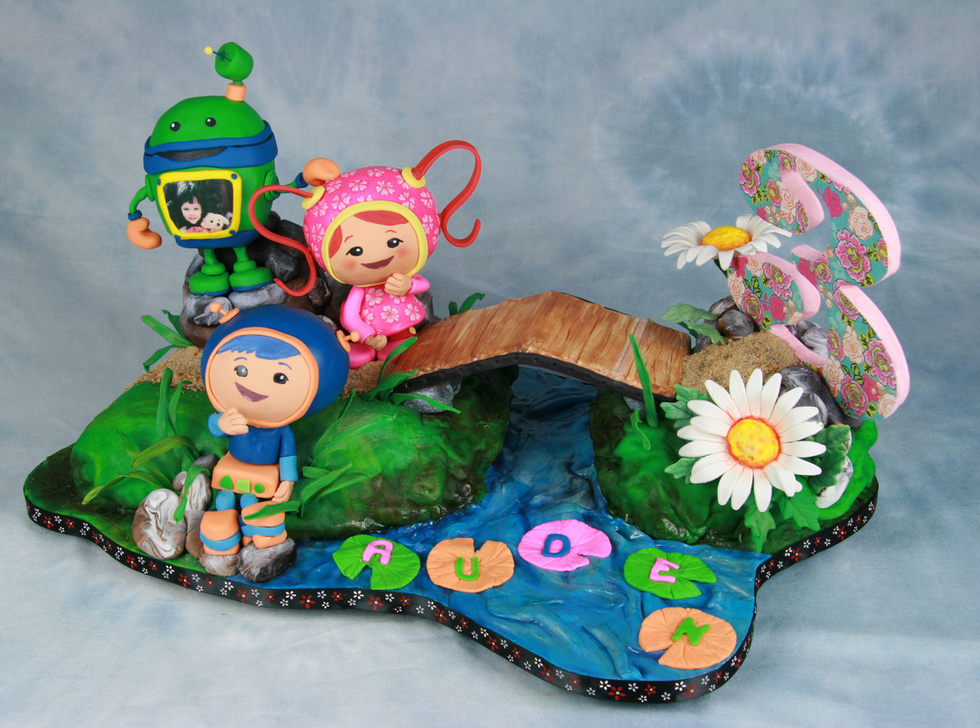A detailed fondant cake topper features Teletubbies characters, a small wooden bridge, and a large floral number 3, all set on a green landscape with a blue stream spelling out the name "AUDREY" on lily pads.