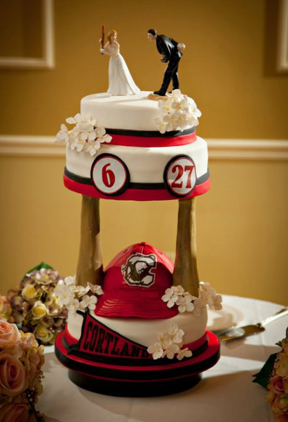 A three-tiered wedding cake decorated in red, black, and white features a baseball theme, including a Cortland Red Dragons cap at the base, jersey numbers 6 and 27 on the middle tier, and a bride and groom figurine on top.
