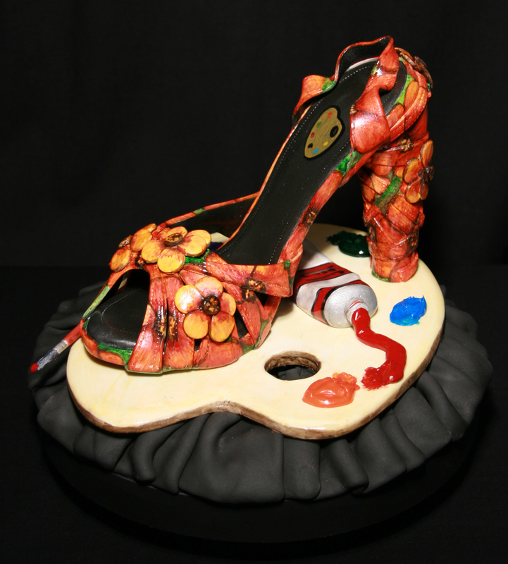 A sculpted high-heeled shoe cake decorated with orange flowers and green leaves sits on a fondant artist's palette base featuring a tube of red paint and blue paint blobs.