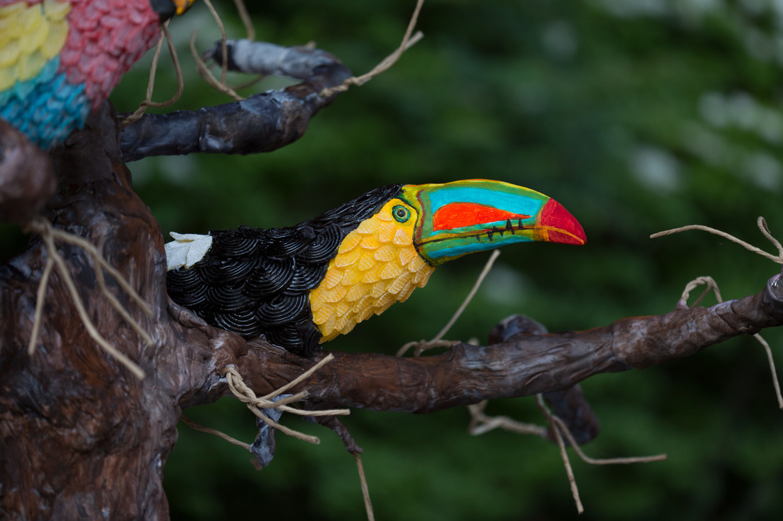 A colorful sugar toucan figurine with a vibrant rainbow beak, yellow chest, and black wings perches on a textured brown branch adorned with twine.