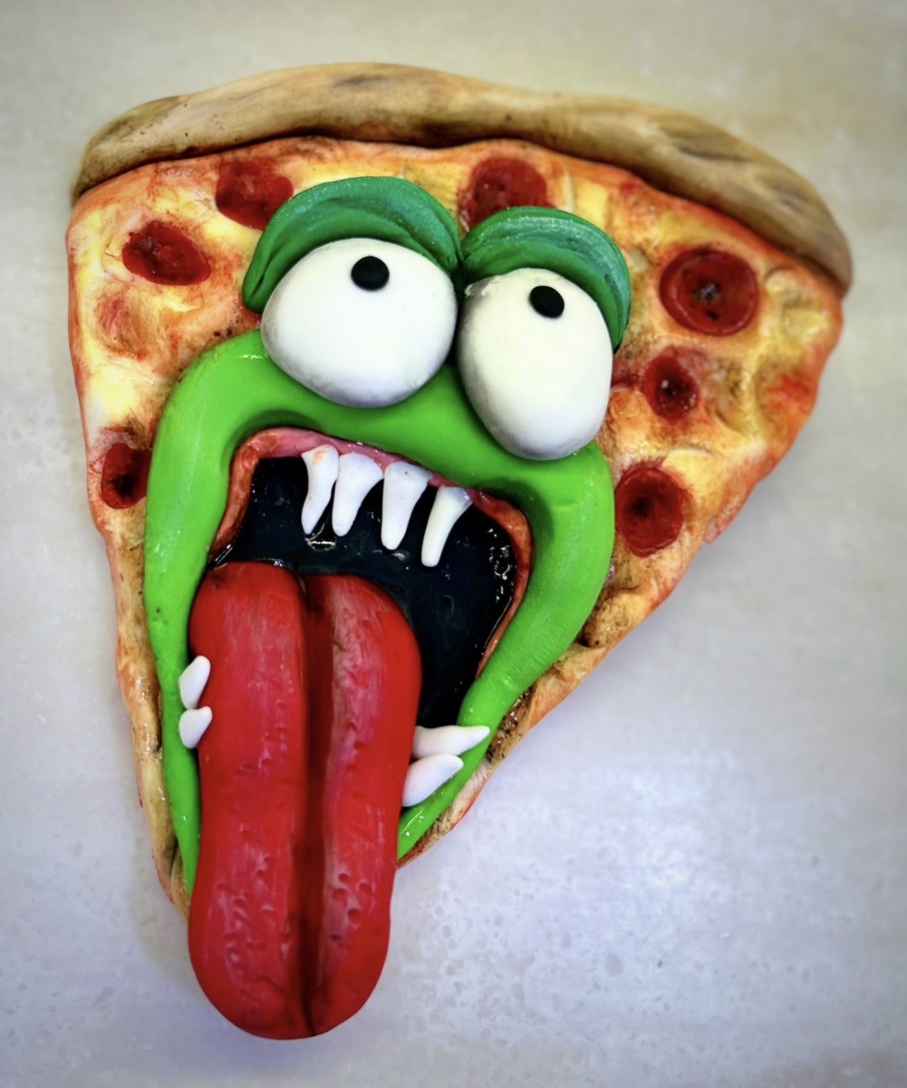 A whimsical cake slice designed as a monstrous pepperoni pizza with a sculpted green monster face made from fondant, featuring bulging white eyes with black pupils, thick green eyebrows, a wide screaming grin revealing sharp white fangs, a long textured red tongue, and the pepperoni slices strategically placed around the face.