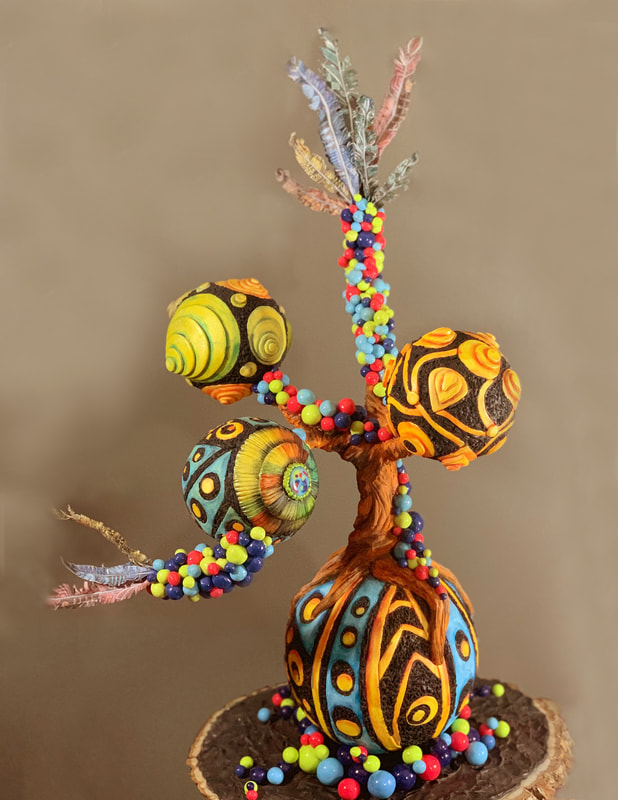 A whimsical, handcrafted cake sculpture resembling a fantastical tree, featuring a bulbous base and branches adorned with colorful beads, patterned spheres, and delicate feathers, all mounted on a rustic wooden slice.