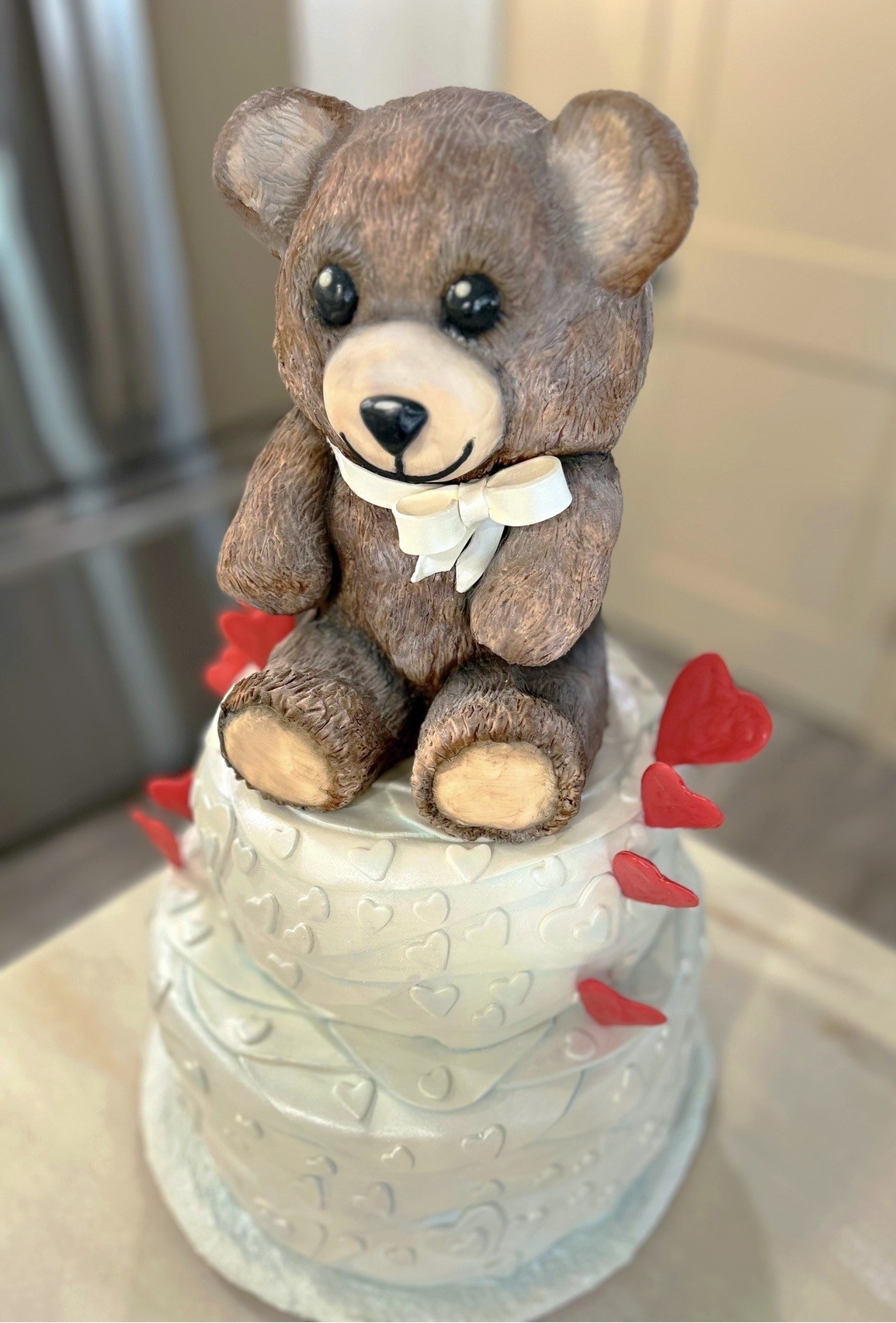 A two-tiered white cake with a sculpted teddy bear topper wearing a delicate white ribbon, the bear having soft brown fur texture and expressive black eyes, the cake decorated with subtle heart patterns embossed into the fondant and vibrant red heart decorations scattered around the base.