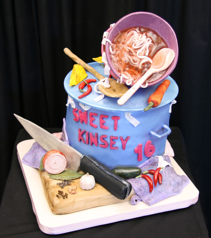 A blue fondant cake shaped like a cooking pot features the name "Sweet Kinsey" and the number "16," topped with a tilted bowl of noodles and surrounded by edible fondant vegetables and a large knife on a cutting board.