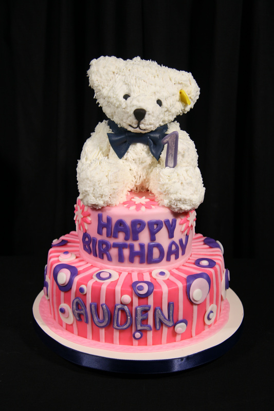A two-tiered pink and purple birthday cake topped with a white teddy bear wearing a blue bow tie and a number one candle.