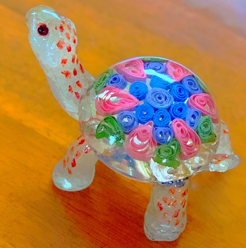 A clear sugar glass turtle figurine with red speckles on its limbs and a shell filled with colorful rolled sugar quilling in pink, blue, and green.