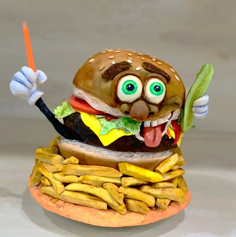 A whimsical sculpture of an anthropomorphic hamburger with googly eyes and a tongue sticking out, holding a french fry and a pickle while sitting atop a pile of fries.