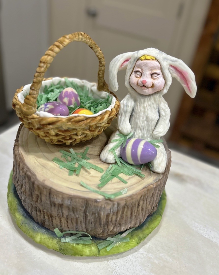 An Easter-themed cake designed as a rustic tree stump with textured bark and light-colored wood grain, topped by a hand-sculpted child in a fluffy white bunny costume with long drooping pink-lined ears and sweet closed-eye smile, beside a wicker basket filled with green shredded paper grass and decorated Easter eggs in purple, yellow, and orange.