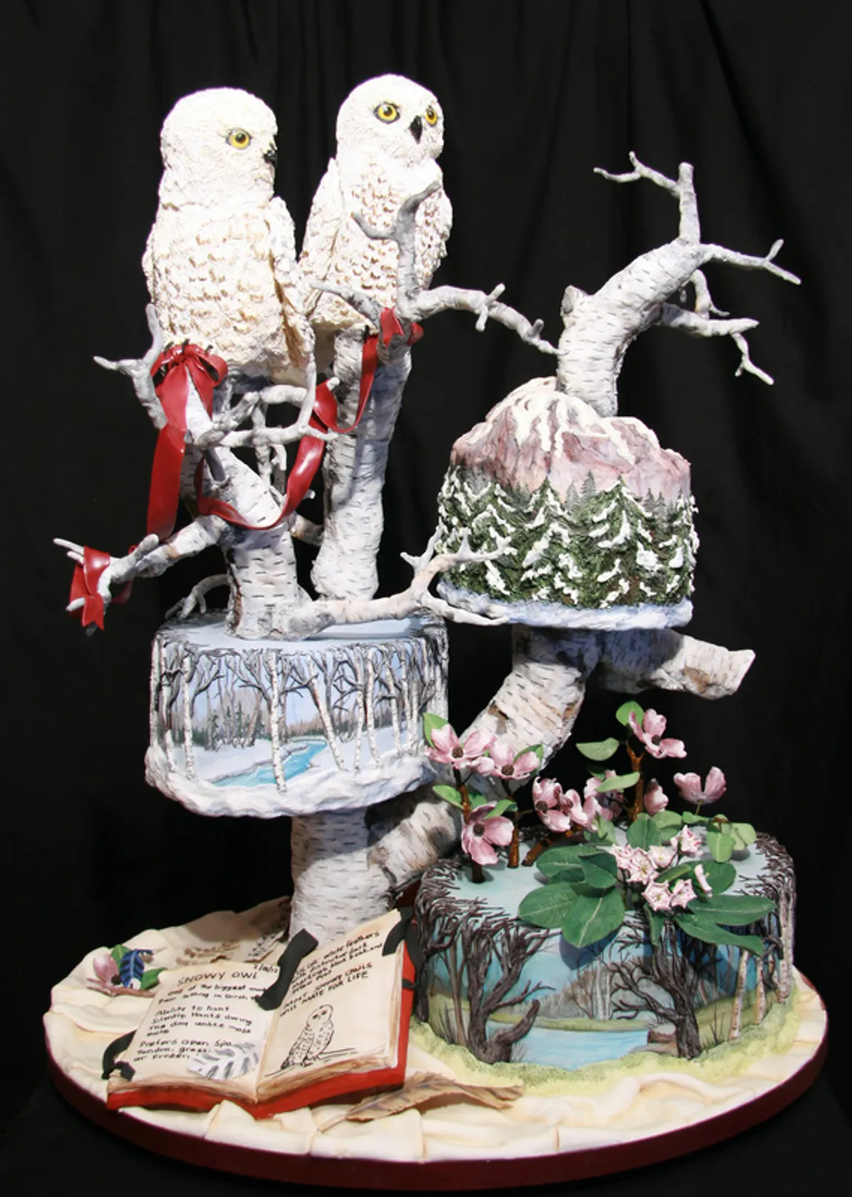 An elaborate, multi-tiered cake decorated with a winter theme features two white owls perched on birch tree branches, surrounded by snowy landscapes, pink flowers, and a miniature open book.