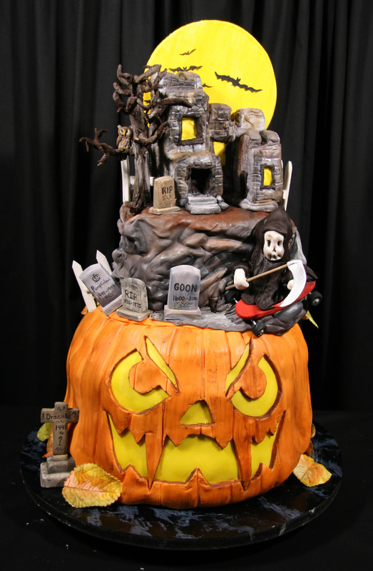 An elaborate Halloween cake features a large orange jack-o'-lantern base topped with a haunted graveyard scene, including a grim reaper, tombstones, and a spooky castle set against a yellow full moon.