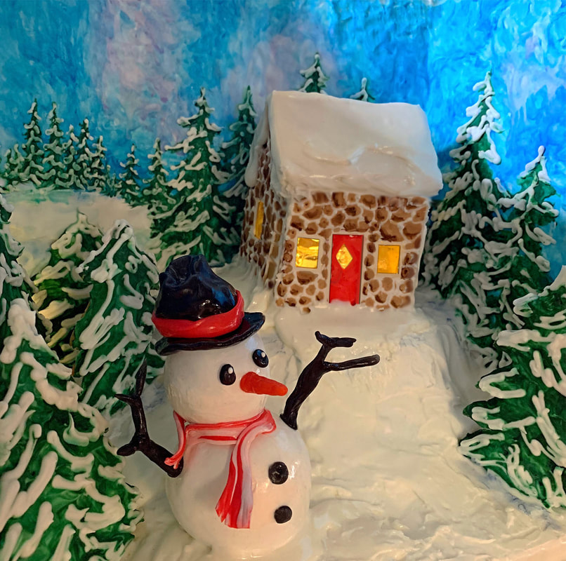 A sculpted cake diorama features a snowman with a top hat and scarf standing before a stone cottage with a red door, surrounded by snowy pine trees against a blue backdrop.