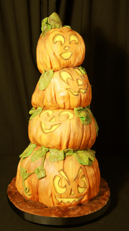 A four-tiered cake sculpted to resemble stacked jack-o'-lanterns, each with a unique carved face and green fondant leaves, sits on a textured brown base against a black background.