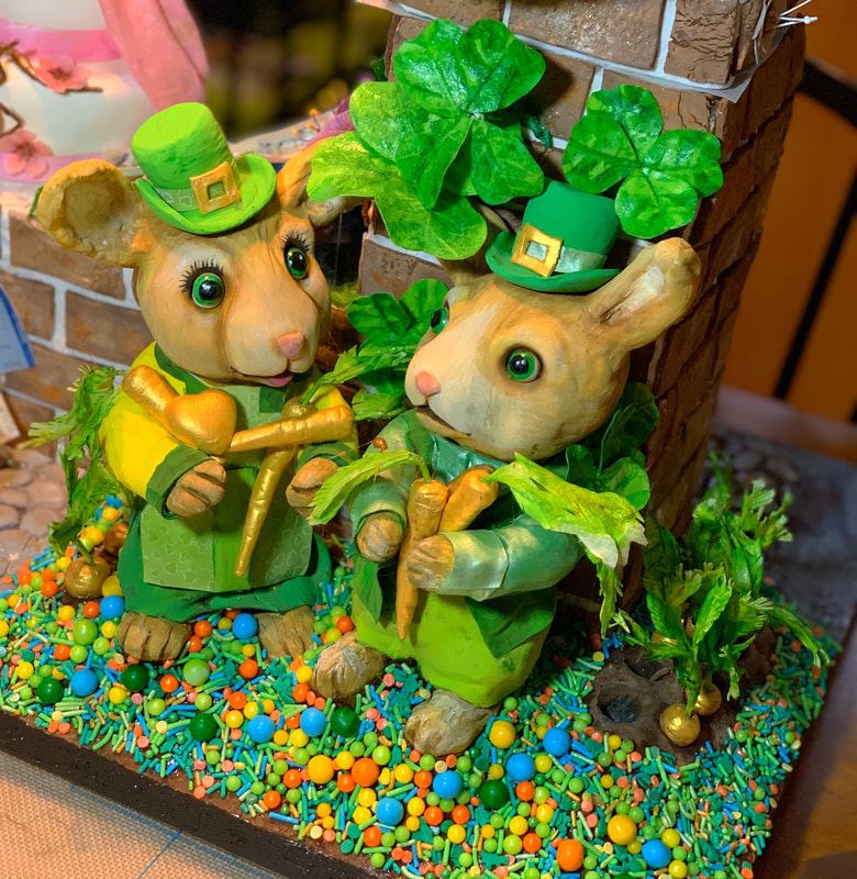 Two whimsical bunny figurines dressed in green suits and leprechaun hats stand amidst colorful sprinkles and shamrocks, holding golden carrots as part of a festive St. Patrick's Day cake topper.