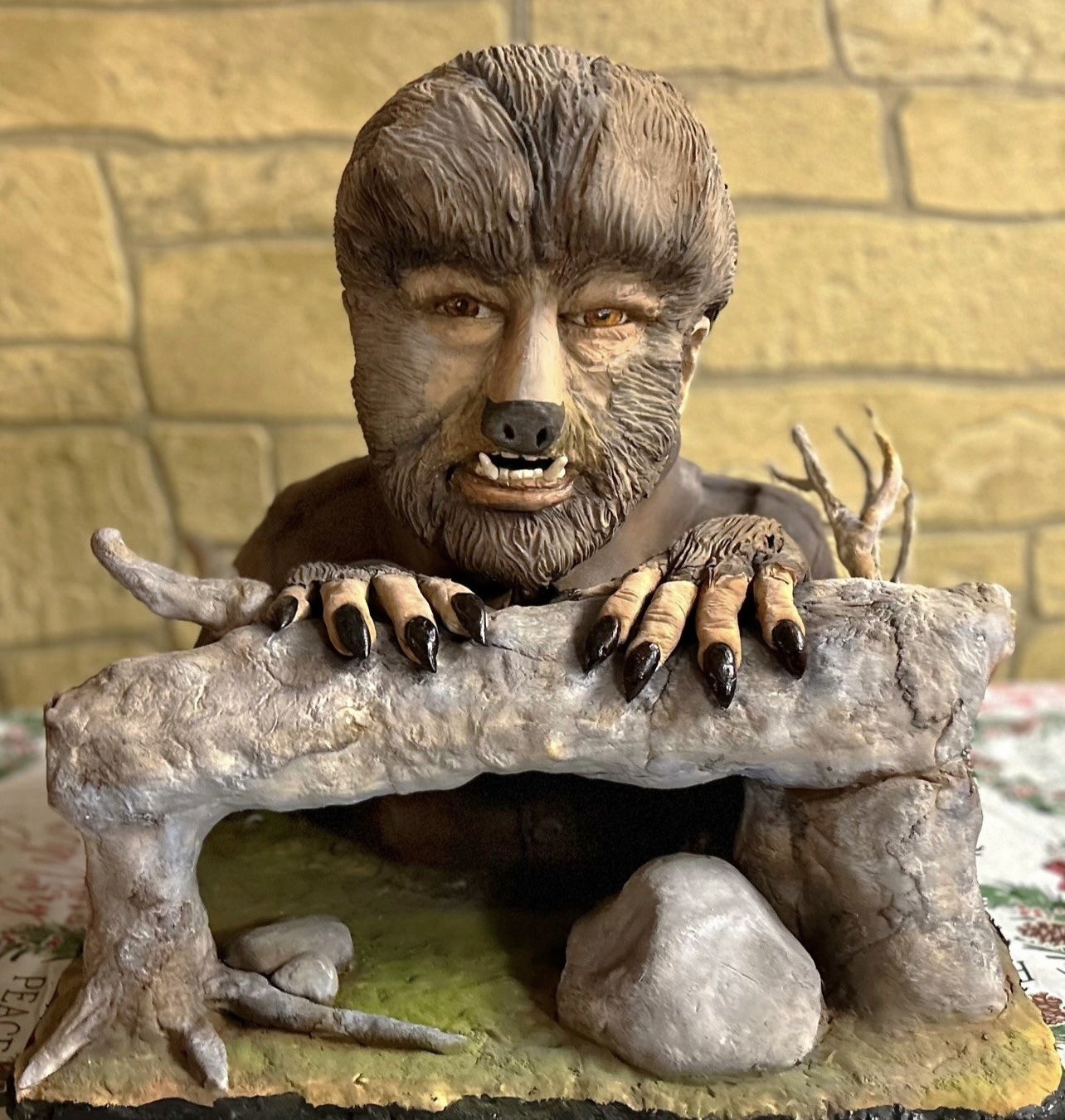 A sculpted cake figurine of a werewolf emerging from a rocky cave-like structure, with fur-covered cheeks, pronounced snout with sharp fangs, intense amber eyes, clawed hands gripping the stone archway, set on mossy green flooring with scattered gray stones and gnarled roots.