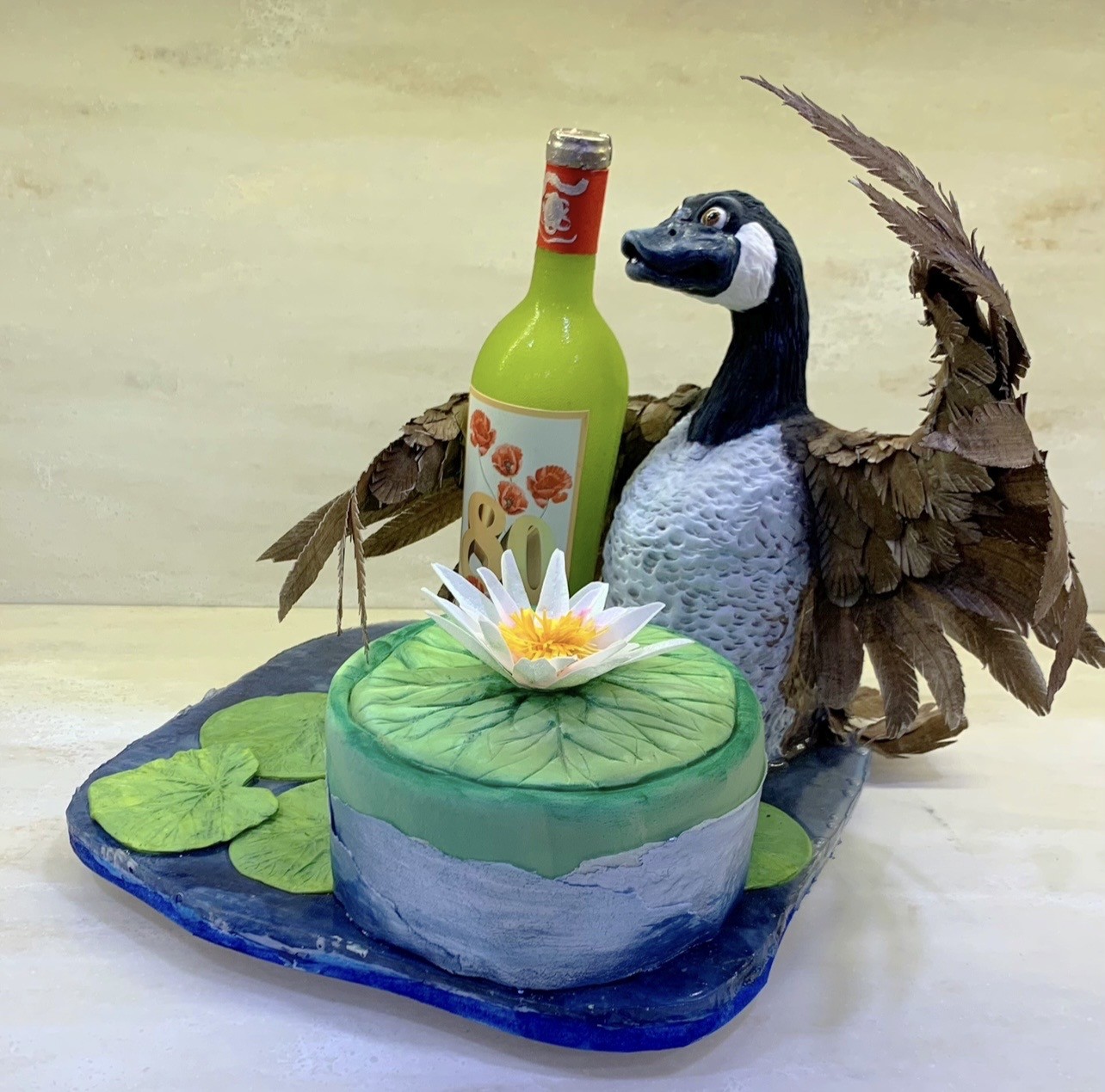 A pond-themed sculpted cake featuring a realistic goose crafted from fondant with layered brown feathers, black head, white cheek patch, wings partially spread as cradling a bright lime-green wine bottle labeled "80" with red foil cap and orange poppy design, round cake base mimicking water with gradient blue frosting, large green lily pad, white water lily, scattered fondant lily pads.