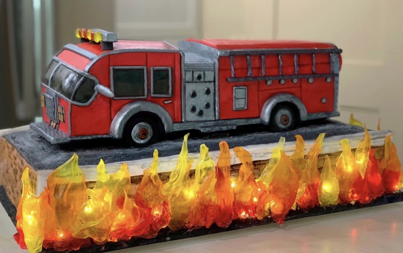 An elaborate fire truck cake sculpted to resemble a classic red fire engine with silver accents on windows, wheels, and structural elements, yellow lights on top, set on a rectangular base mimicking asphalt with vibrant translucent flames made from edible gelatin or sugar glass illuminated from within.