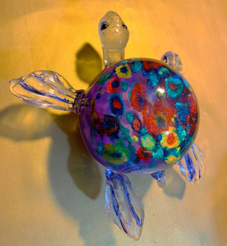 A small sugar figurine of a sea turtle features a translucent body with blue-tinted flippers and a vibrant, multicolored shell decorated with purple, red, green, and blue speckles.