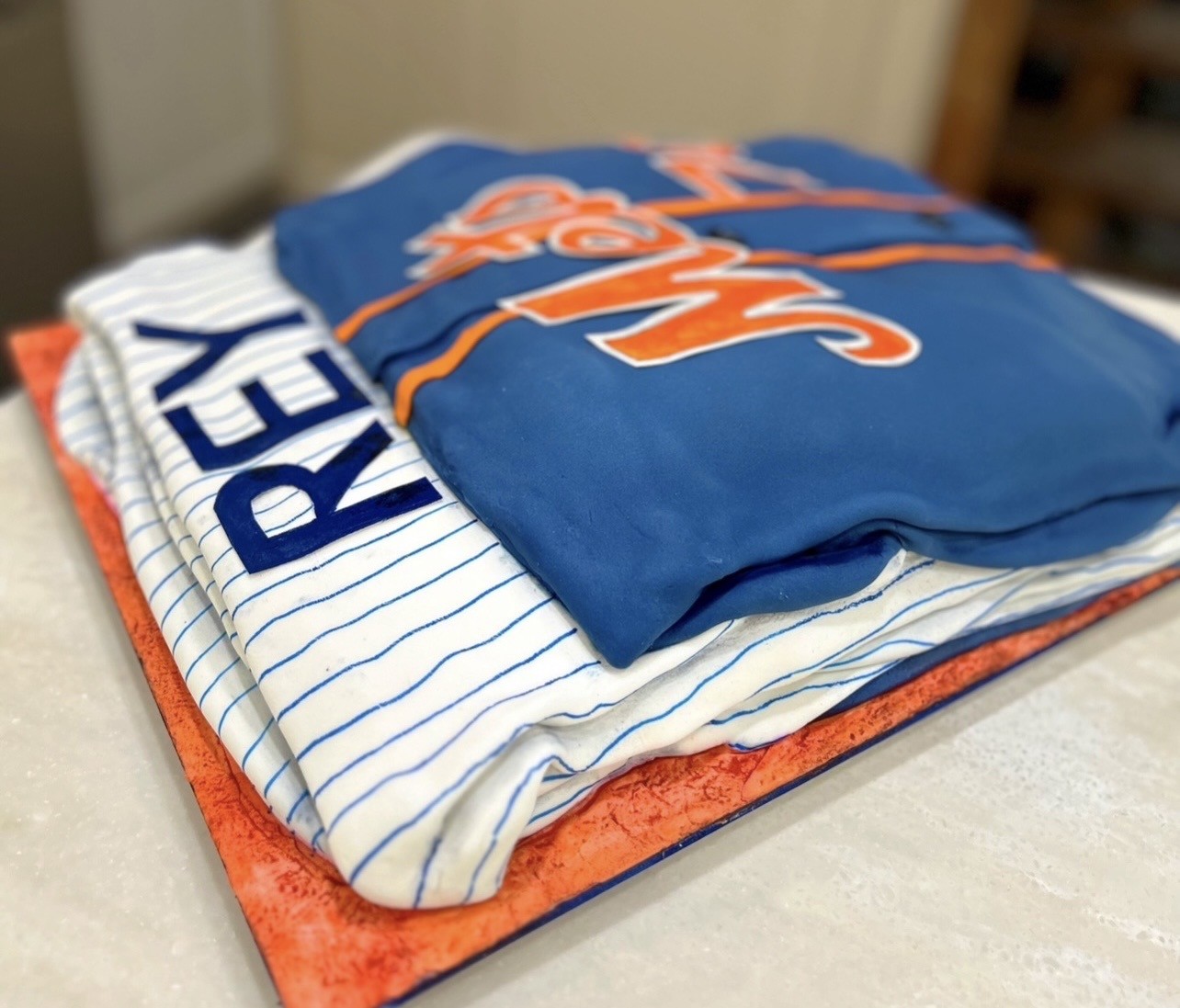 A cake designed to resemble two folded New York Mets baseball jerseys on an orange textured board — the top jersey in royal blue fondant with bold orange and white "Mets" script, beneath it a white pinstriped jersey with the name "REY" in navy-blue block letters.